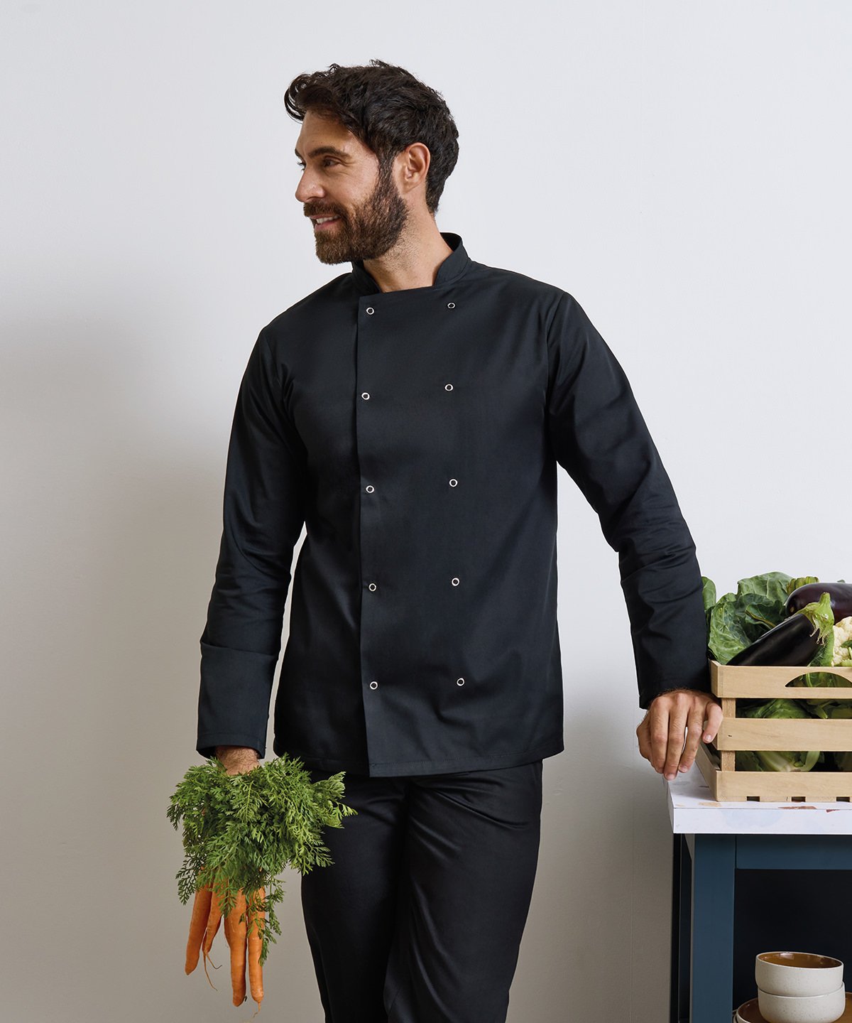 Man in a black chef's coat holding vegetables and a crate of greens against a neutral background